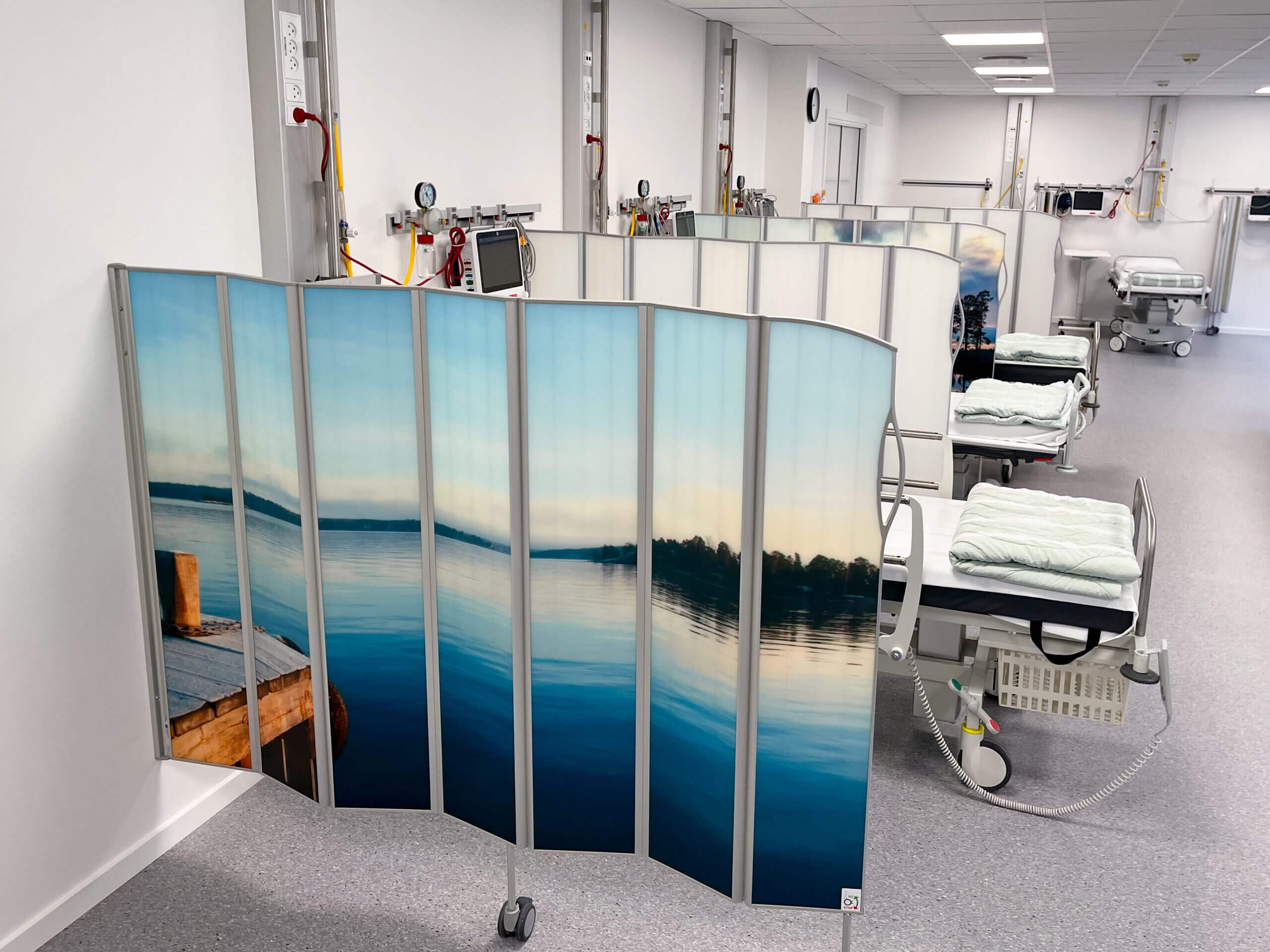 Böjd Silentia vikskärm med en lugn sjö i PhotoPanels™ som avdelar en sjukhussal, illustrerar en kundberättelse.