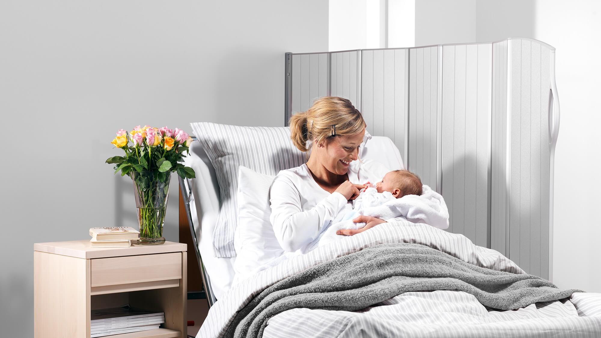 Äiti pitelee vastasyntynyttään sairaalasängyssä läpikuultavan Daylight-sermin vieressä, joka päästää lävitse pehmeää valoa.