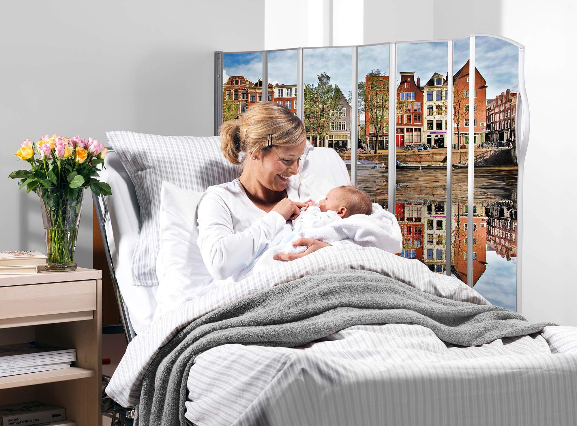 Mother holding her baby in bed beside a Silentia screen printed with the Amsterdam PhotoPanels™ canal view.
