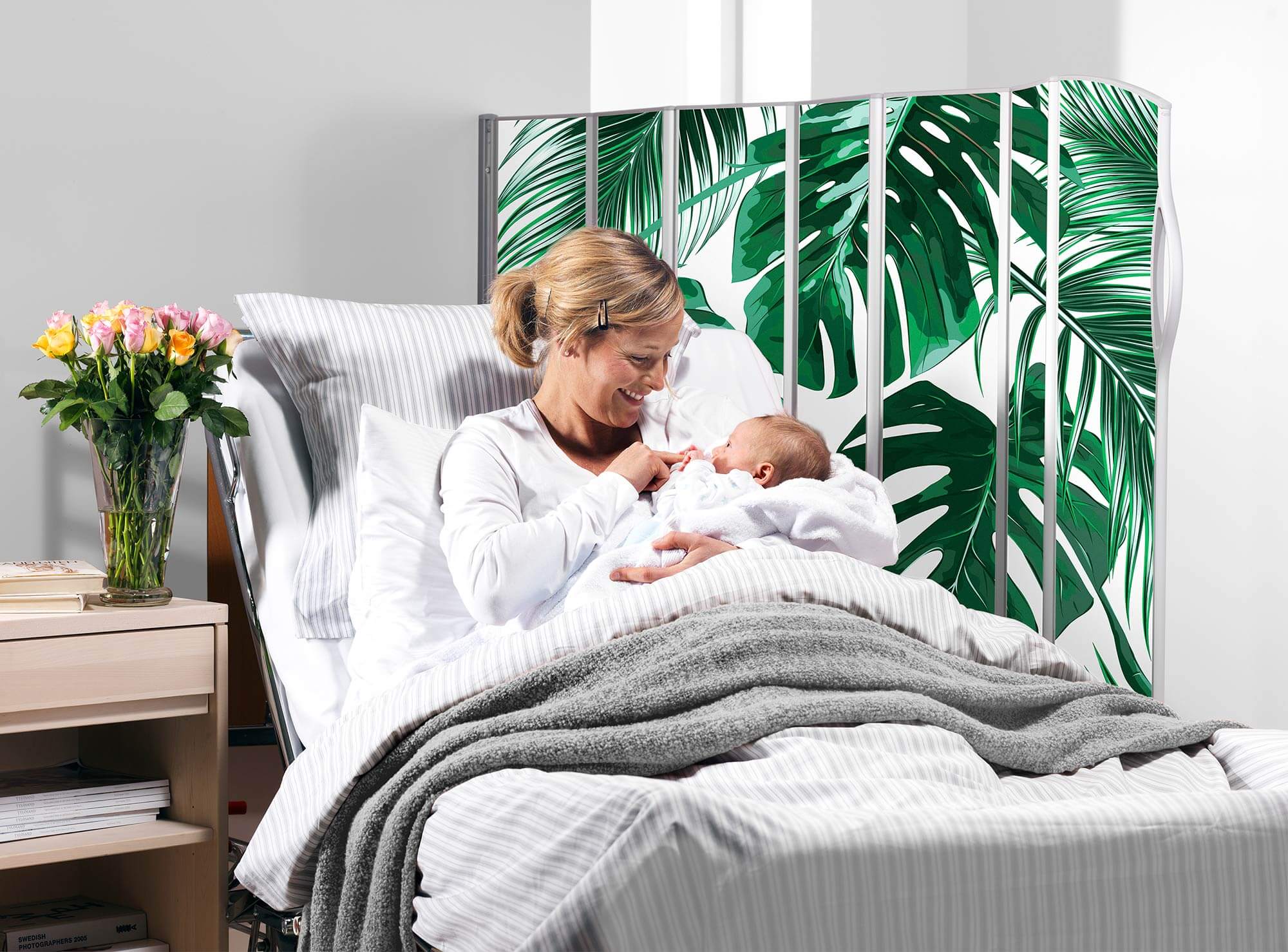Mother holding her baby in bed beside a Silentia screen printed with the Tropical palm leaves PhotoPanels™ image.