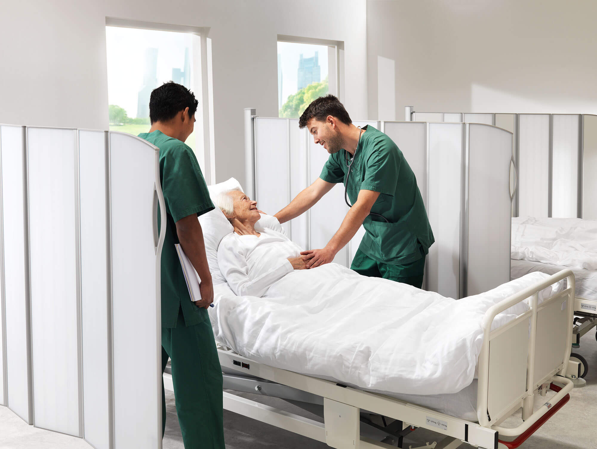 Doctor with stethoscope examining an elderly patient in a bed enclosed by Silentia folding screens.
