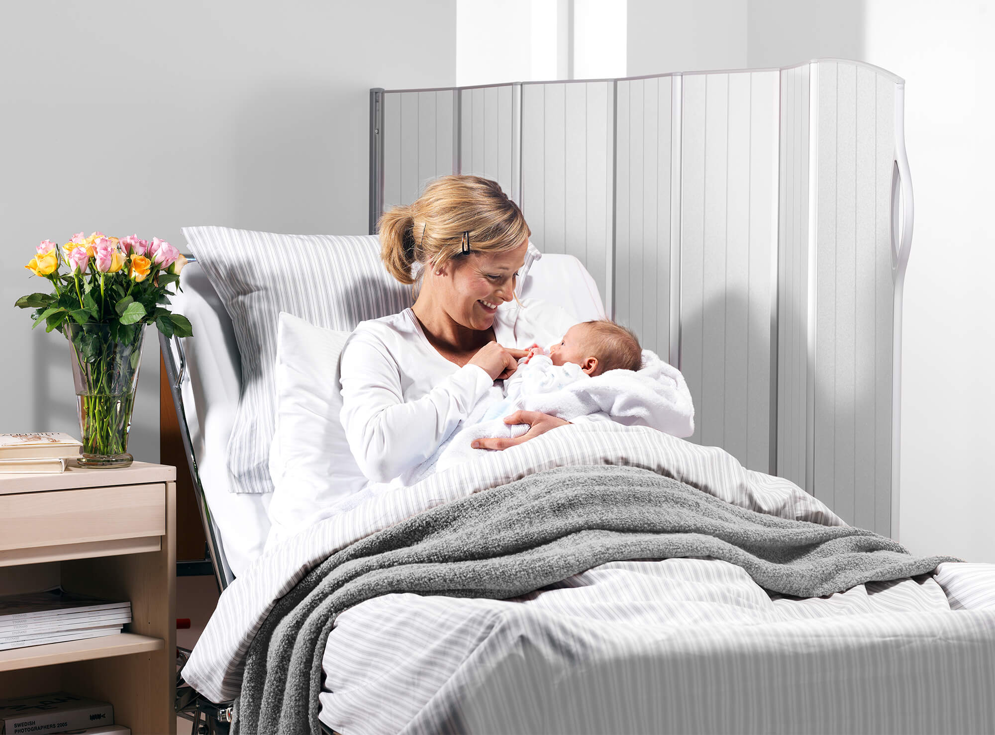 Mother holding her newborn in a hospital bed beside a translucent Daylight screen letting in soft light.