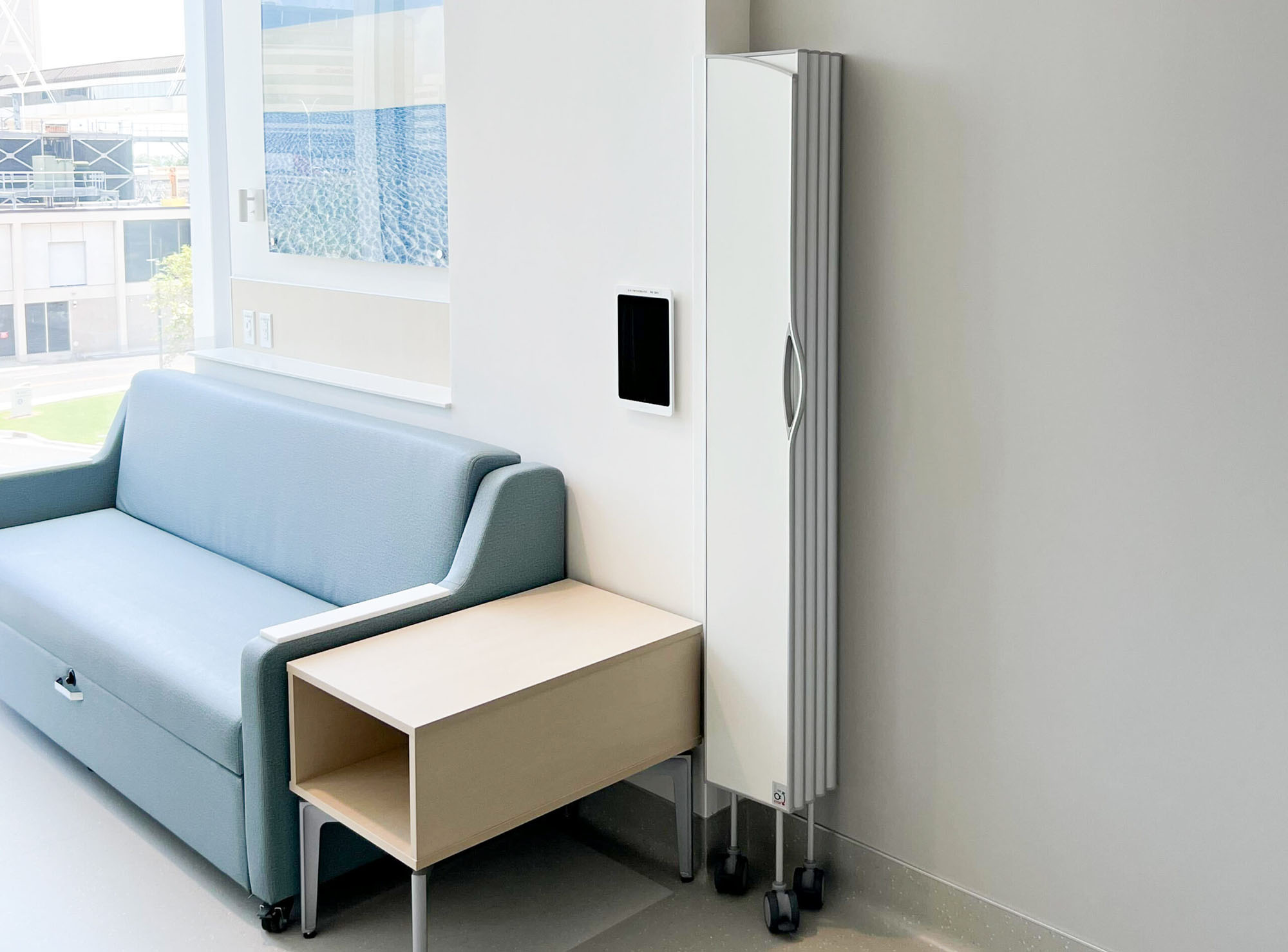 White Silentia Fold screen folded neatly in the corner of a calm hospital room beside a sofa.