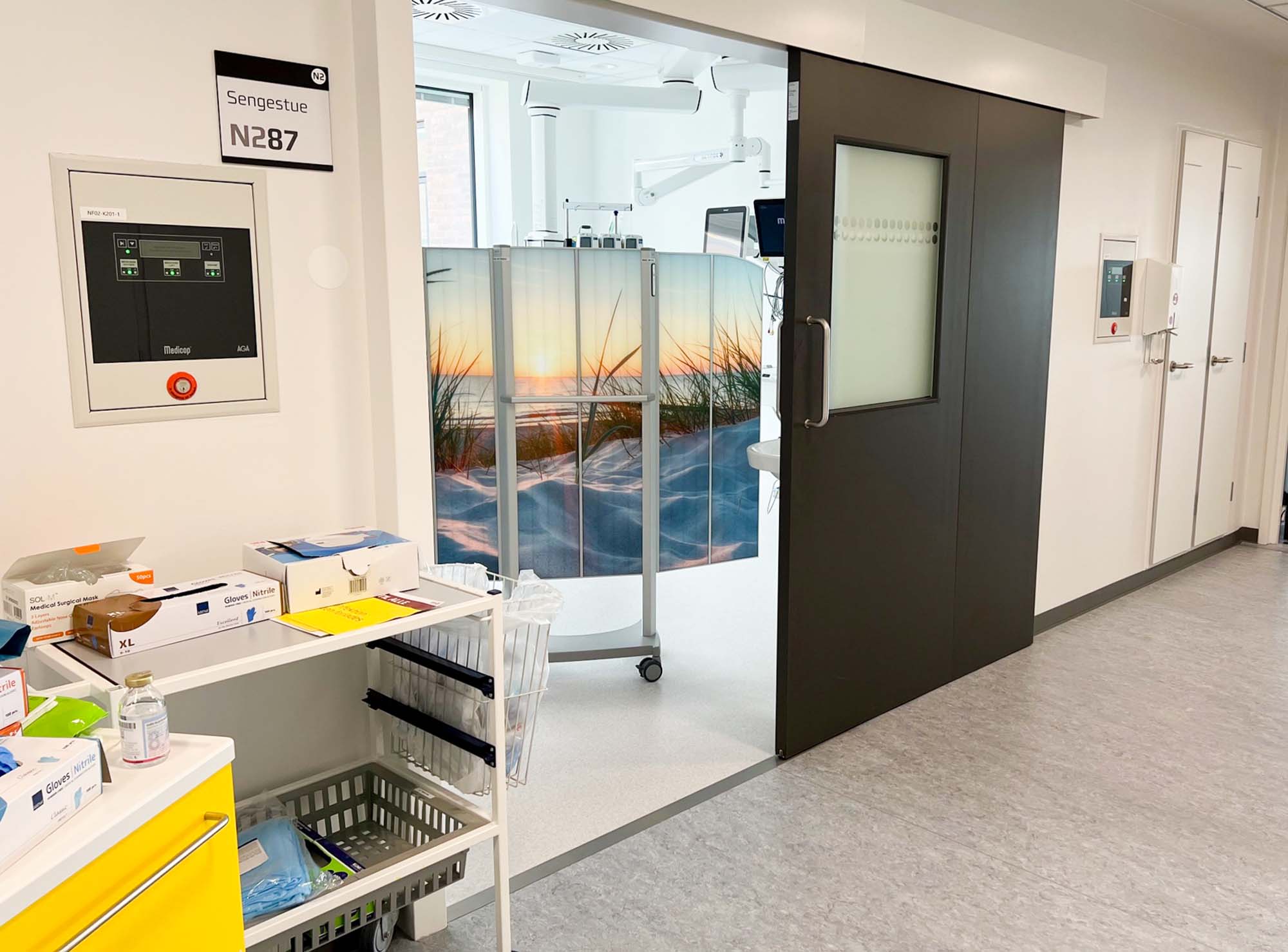 Silentia Fold screen with beach sunset PhotoPanels™ shielding a monitored-care room seen from a corridor doorway.