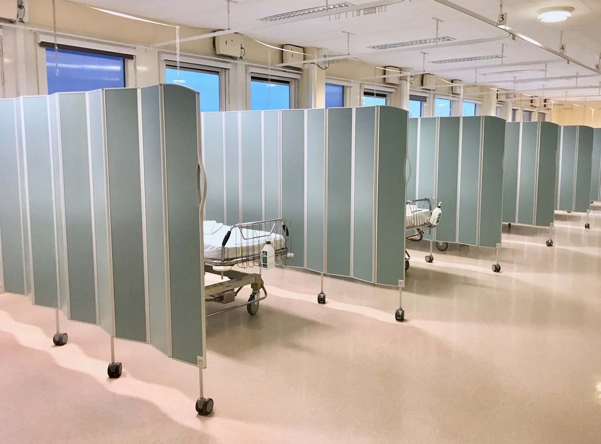 Green-gray Silentia Fold screens partitioning a row of beds in a large open ward by windows.
