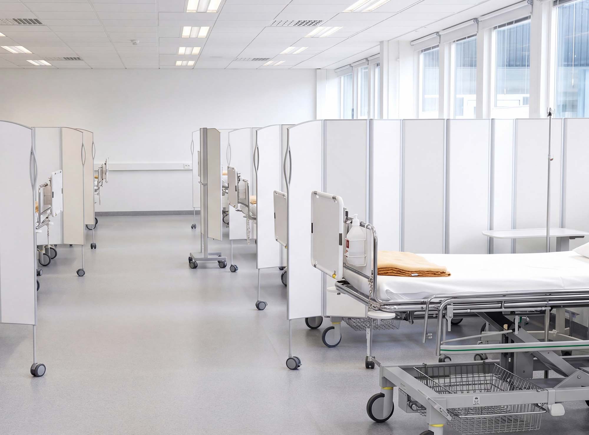 White Silentia Fold screens separating a long row of beds in an open care space by tall windows.