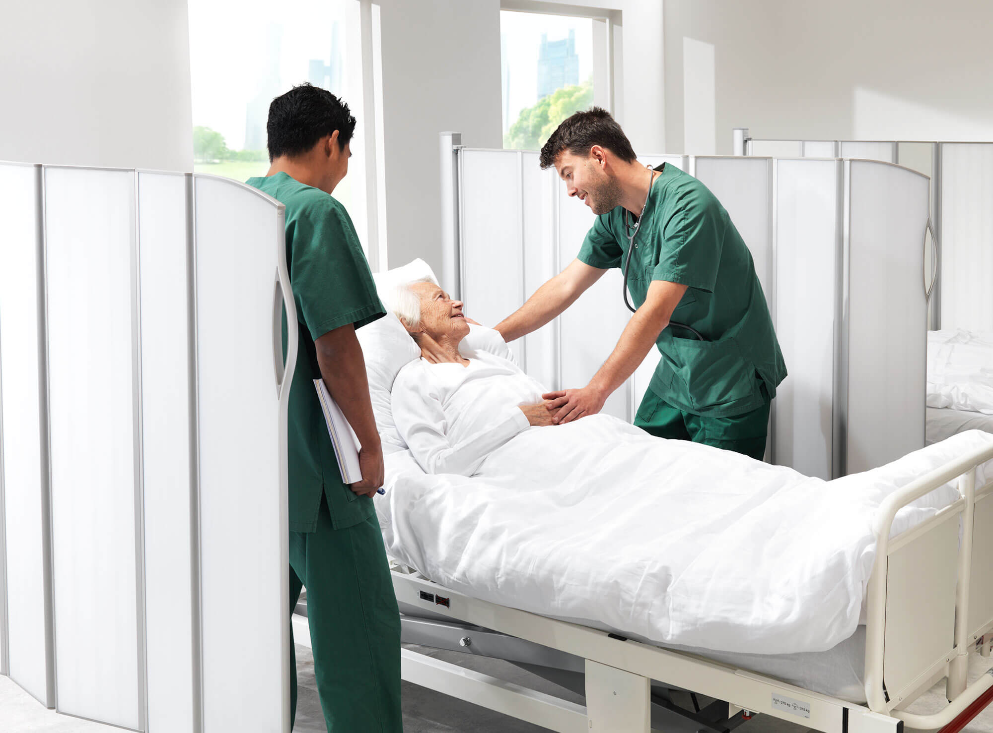Zwei Pflegekräfte in grüner Kleidung versorgen einen älteren Patienten in einem Krankenbett zwischen weißen Silentia-Schirmen.