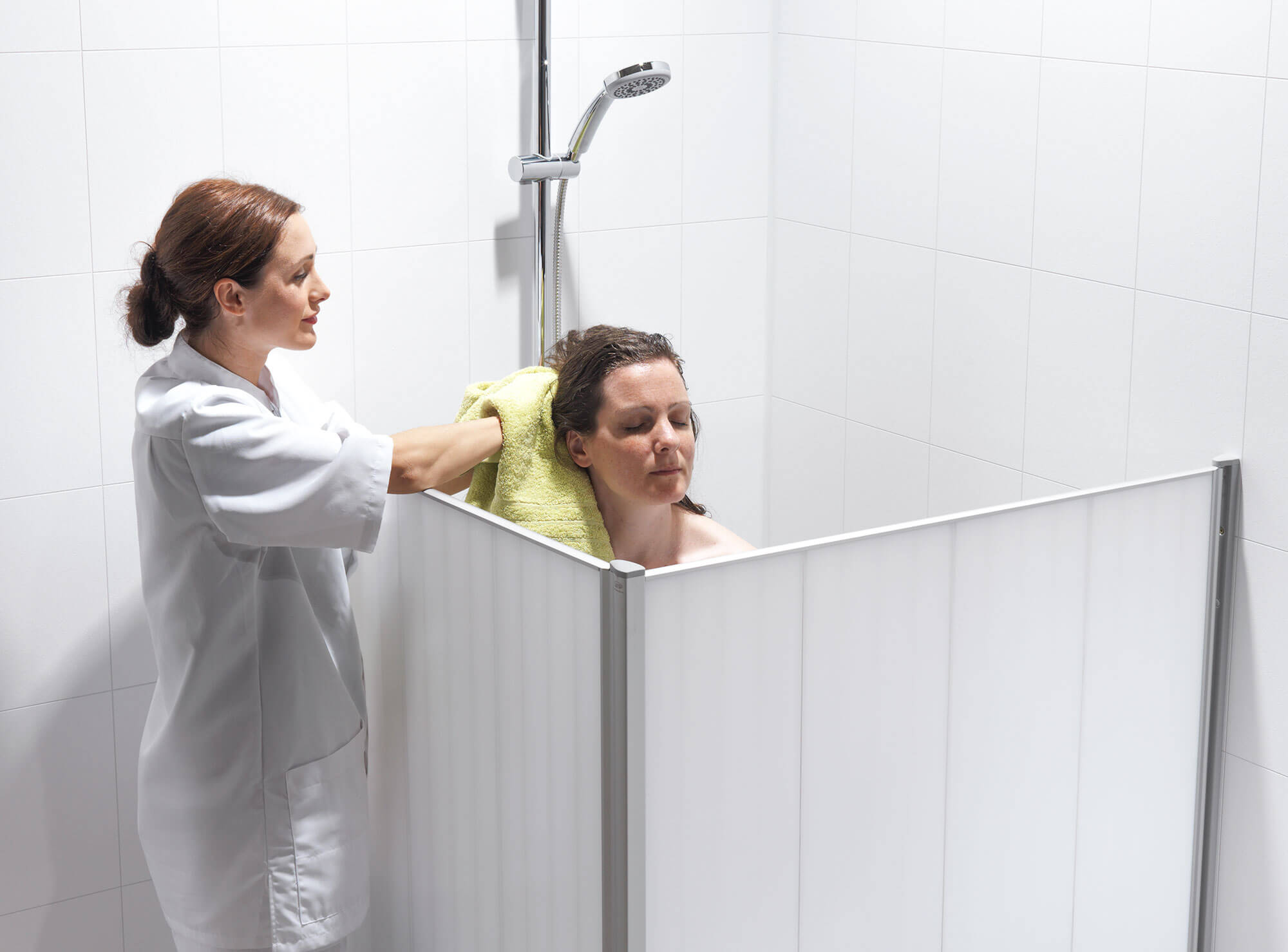 Betreuerin trocknet die Haare einer Frau, während diese hinter weißen Silentia Shower-Wandpaneelen in einem gefliesten Bad duscht.