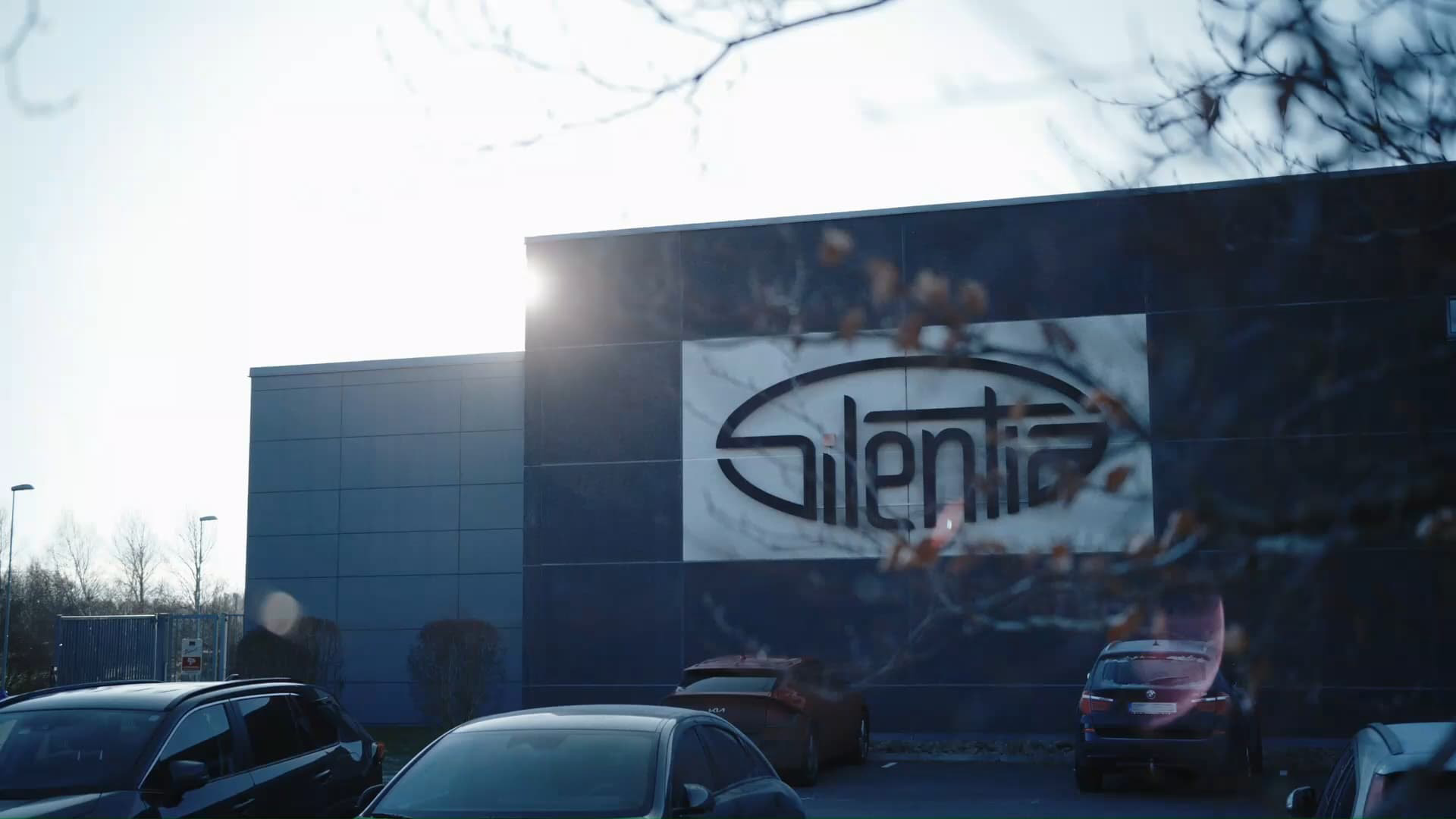 Exterieur van het hoofdkantoor van Silentia met het bedrijfslogo op de donkere gevel, auto's geparkeerd ervoor en kale takken op de voorgrond.