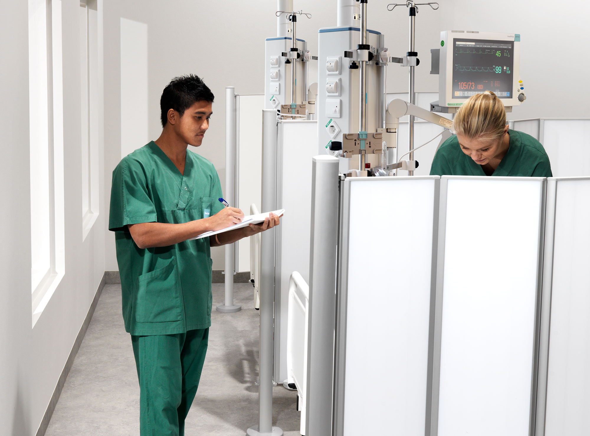 Två kliniker i gröna skrubbar som arbetar vid en patientövervakningsstation avdelad med vita Silentia-skärmar i en sjukhusplats.