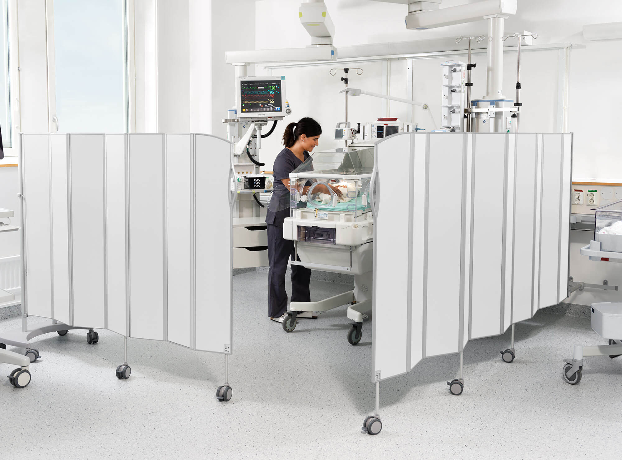 Sjuksköterska som sköter ett nyfött barn i en kuvös på en neonatalplats omgiven av vita mobila Silentia-skärmar.